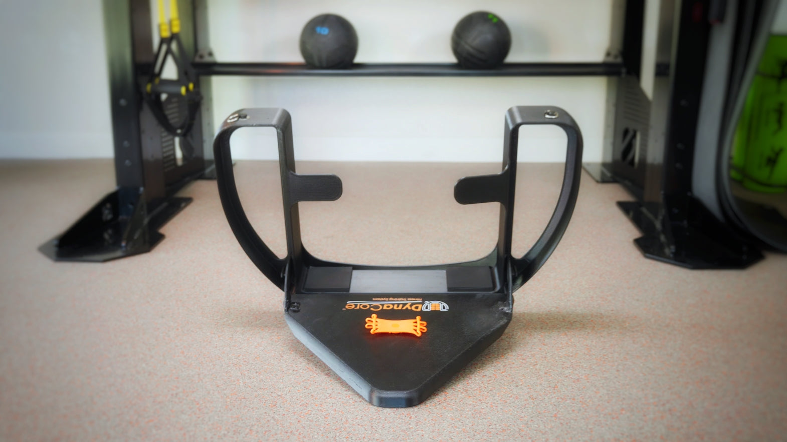 A black and orange DynaCore Basic Fitness Trainer positioned on a gym floor, with exercise balls and equipment in the background.